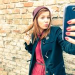 56299814 - young pretty woman taking selfie outdoors - female winter fashion portrait - teenager student holding mobile phone for selfi photo next to brick wall background - soft and hazy vintage filtered look